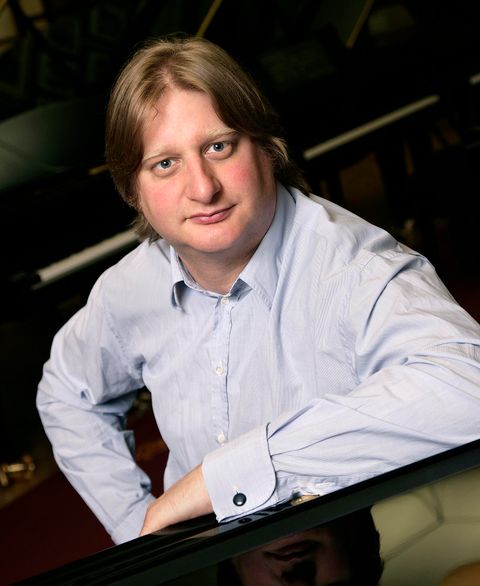 Ein Mann sitzt am Klavier und schaut in die Kamera. Er trägt ein hellblaues Hemd und hat blonde Haare.