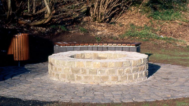 Round stone fountain with bench and trash can in the background, surrounded by trees.