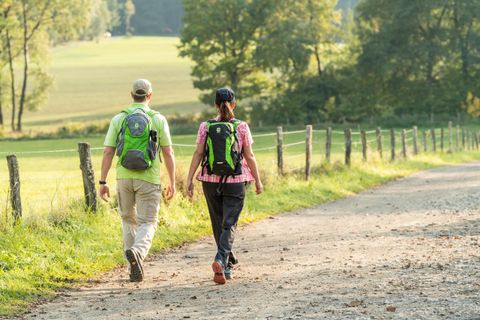 Un couple marche sur un chemin de campagne, entouré d'arbres et de prairies. Tous les deux portent des sacs à dos et profitent de la nature.