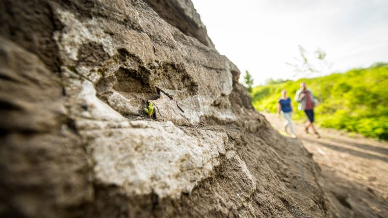 Massive Gesteinsschichten im Vordergrund, an der zwei Wanderer vorbeilaufen.