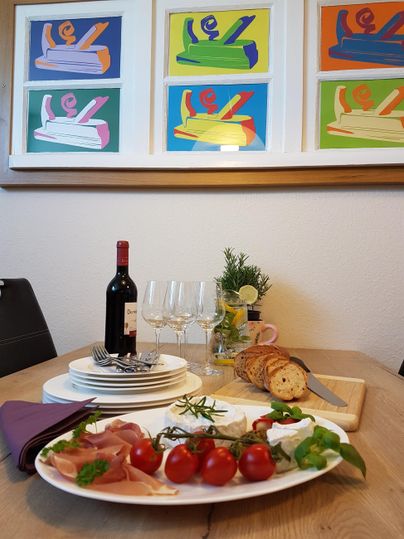 Ein schön gedeckter Tisch mit einem Teller voller Tomaten, Prosciutto und einer flaschen Rotwein. Im Hintergrund hängen bunte Kunstwerke an der Wand.
