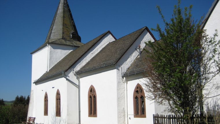 Weiße Kirche mit spitzem Turm und gotischen Fenstern vor blauem Himmel.