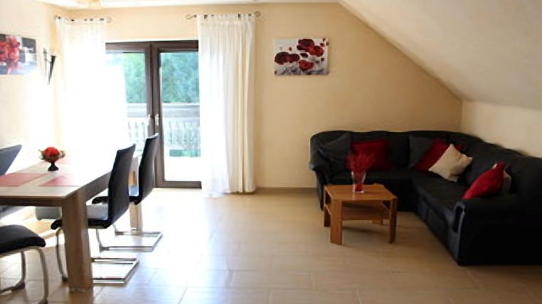 Ein modernes Wohnzimmer mit einem gemütlichen Sofa, einem großen TV  und einer Essgruppe,  Durch die Balkontüren zum Ost-Balkon flutet das Licht der aufgehenden Sonne in den Raum und und verleiht eine helle Atmosphäre.