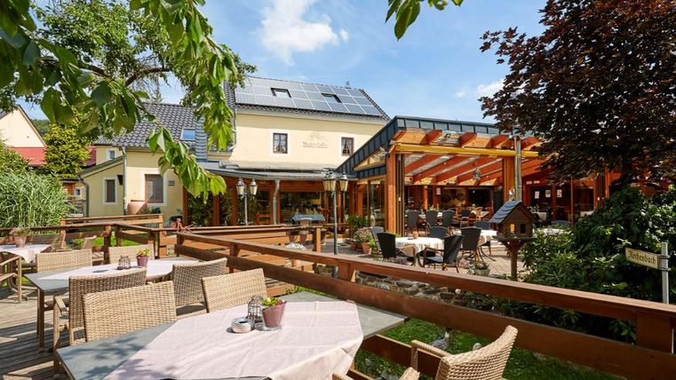 Ein einladendes Restaurant mit einer gemütlichen Terrasse und Holzmöbeln. Die Umgebung ist grün und sonnig, ideal für eine entspannte Mahlzeit im Freien.