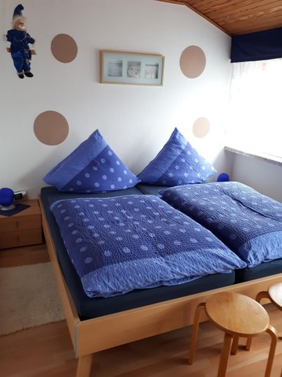 Ein gemütliches Schlafzimmer mit einem großen Doppelbett und blauen Bettwaren. Kleine Holzstühle und eine helle Atmosphäre sorgen für ein einladendes Ambiente.