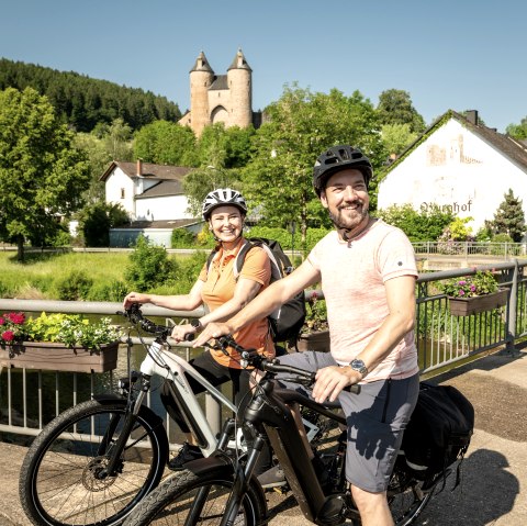 A man and a woman on bicycles stop on a bridge in M&uuml;rlenbach. Behind them, you can see the Kyll river, several houses and Bertrada Castle.