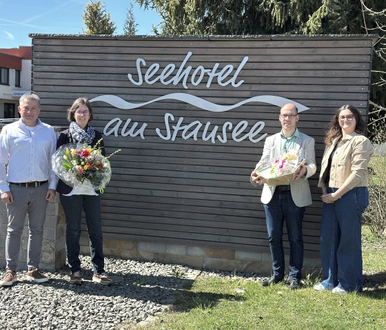 Vier Personen stehen vor einer Holzwand mit der Inschrift &quot;Seehotel am Stausee&quot;. Zwei der Personen halten Blumen und einen Präsentkorb in den Händen.