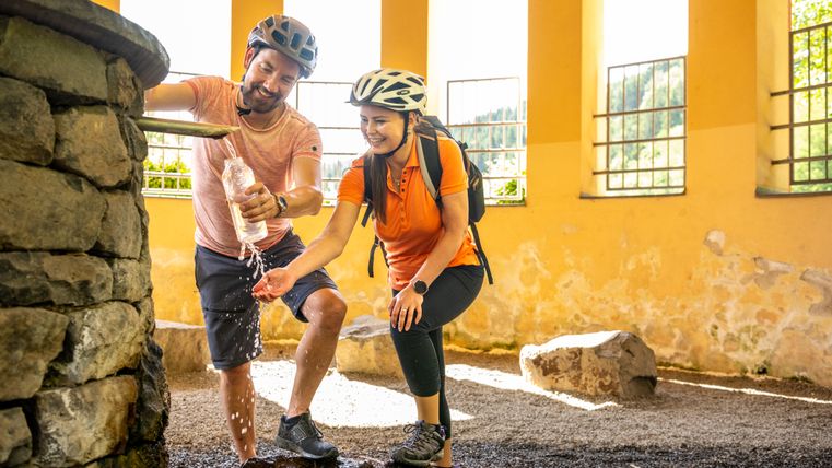 Zwei Personen mit Fahrradhelmen füllen eine Flasche an einer Quelle auf.