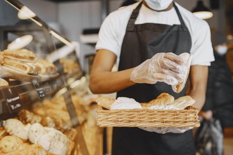 Ein Bäcker mit Schürze und Handschuhen legt frisches Gebäck in einen Korb in einer Bäckerei.