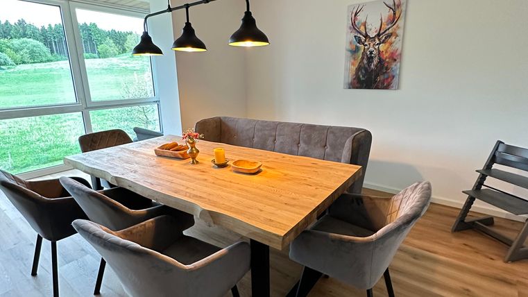 Ein modernes Esszimmer mit einem großen Holztisch und gepolsterten Stühlen. Die großen Fenster bieten einen schönen Blick auf die Landschaft.