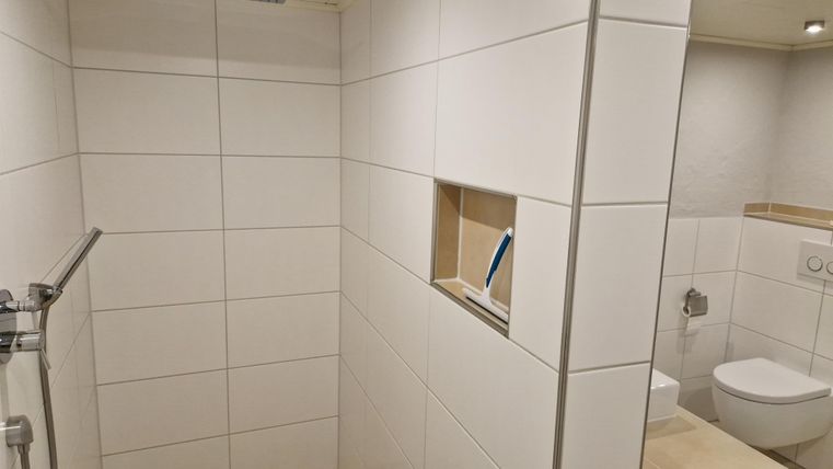 Eine moderne Dusche mit weißen Fliesen und einem klaren Duschbereich. Im Hintergrund ist eine Toilette sichtbar.