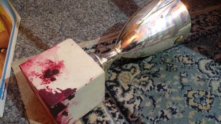 Ein silberner Pokal liegt umgekippt auf einem gemusterten Teppich. Der Sockel hat rote Blutflecken.