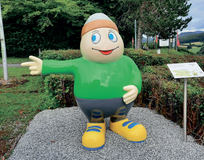 Statue von Maskottchen Willi Basalt mit grünem Shirt, grauer Hose und gelben Schuhen zeigt nach links. Hinter ihm Bäume und ein Parkplatz.