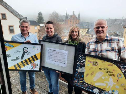 4 Personen stehen auf einem Balkon oberhalb einer Stadt. Sie halten 3 eingerahmte Bilder in den Händen. 2 der Bilder sind von Kindern gebastelte Collagen und das Bild in der Mitte zeigt einen Spendenscheck.