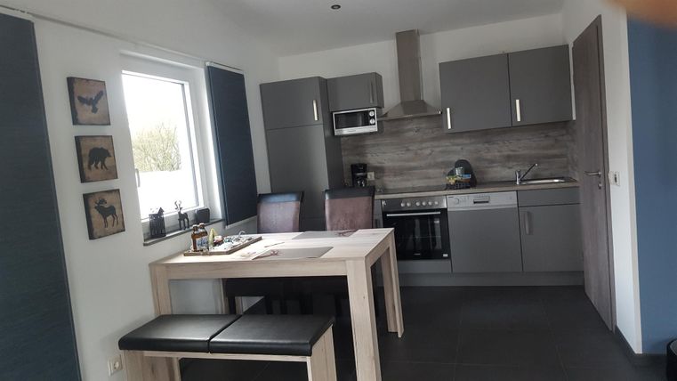 Eine moderne Küche mit grauen Schränken und einem Holzstammtisch. Es gibt Stühle in verschiedenen Farben und eine große Fensterfront, die Licht hereinlässt.