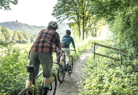 Twee fietsers op gravel fietsen over een smal natuurpad naast een dicht bos.