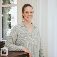 Eine Frau steht hinter einem Tresen und lächelt in die Kamera. Hinter ihr einige Fenster und Regale mit regionalen Produkten.