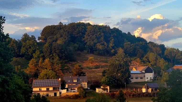 Eine ruhige Landschaft mit einem Hügel im Hintergrund und einigen Häusern im Vordergrund. Die Sonne taucht die Szene in warmes Licht.