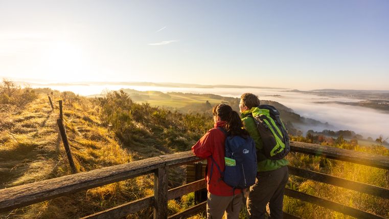 Ein Paar steht an einer Aussichtplattform und blickt auf eine nebelige Landschaft im Morgenlicht. Die Szenerie zeigt sanfte Hügel und einen klaren Himmel.