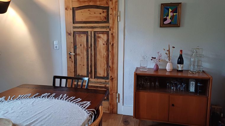 Een gezellige eetruimte met een houten tafel en stoelen. Op de achtergrond is een houten deur en een dressoir met decoratie en drankjes te zien.