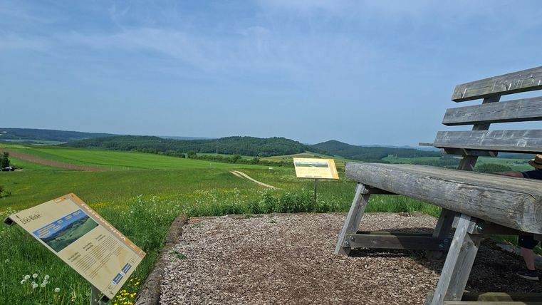 Eine Holzbank steht auf einem Aussichtspunkt mit grünem Feld und blauen Himmel im Hintergrund. Neben der Bank sind Informationstafeln aufgestellt.