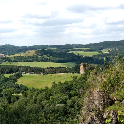 Blick auf eine h&uuml;gelige Wiesen- und Waldlandschaft mit vereinzelten H&auml;usern und Windr&auml;dern dazwischen. Rechts ragen Felsen ins Bild.
