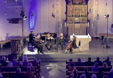 Fünf Musiker an Streichinstrumenten und Klavier musizieren vor dem Kirchenaltar. Davor die Sitzbänke voller Besucher.