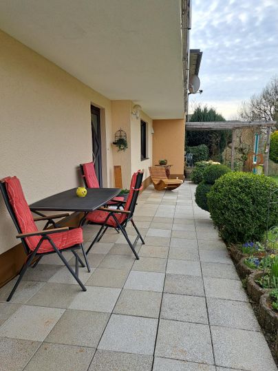 Ein gemütlicher Außenbereich mit einem Tisch und Stühlen auf einer Steinterrasse. Grüne Büsche und Pflanzen verleihen der Umgebung eine einladende Atmosphäre.