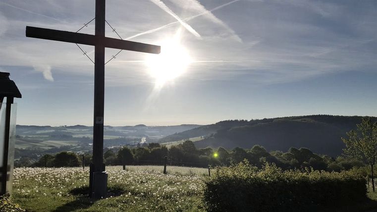Holzkreuz auf einer Wiese mit Blick auf die hügelige Landschaft der Eifel bei Sonnenaufgang.