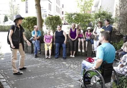 A group of about 11 people, consisting of adults and children, sit next to each other in a circle on a wall next to a street. A group leader stands in front of the group and explains something.