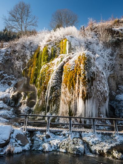 Verschneiter Wasserfall Dreimühlen mit Eiszapfen und Moos, blauer Himmel im Hintergrund.
