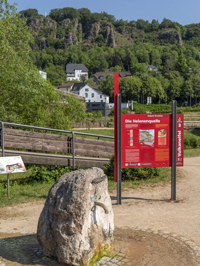 Ein Brunnen mit einem Felsen und einer Infotafel zur Helenenquelle im Vordergrund, umgeben von grüner Landschaft und Hügeln.
