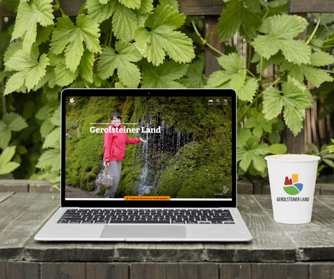 Ein Holztisch draußen vor einer bewachsenen Wand. Auf dem Tisch steht ein Laptop, auf dem die Homepage der Ferienregion Gerolsteiner Land geöffnet ist. Daneben ein Pappbecher mit dem Logo der Touristik GmbH Gerolsteiner Land darauf.
