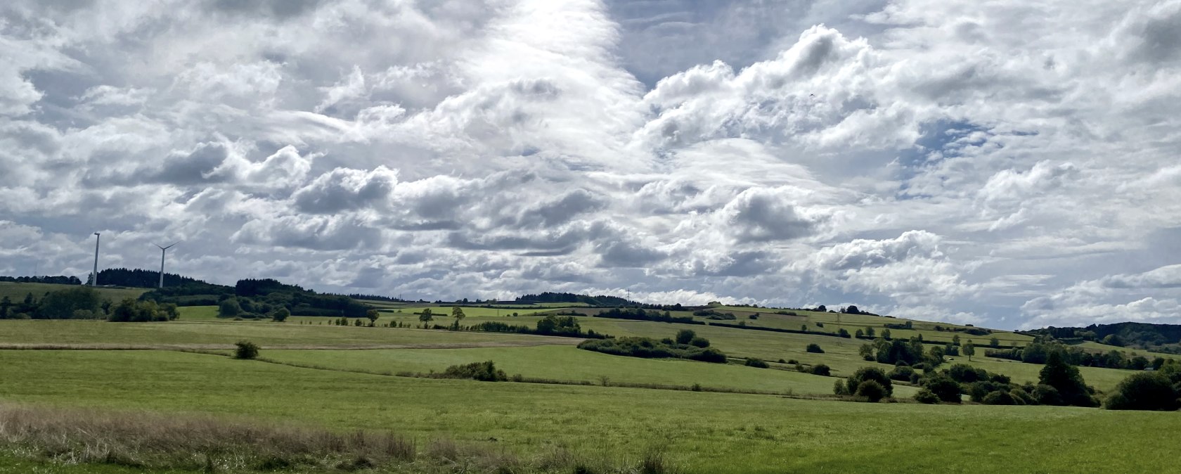 Wide green fields under a cloudy sky, wind turbines on the horizon, paved path in the foreground., &copy; Touristik GmbH Gerolsteiner Land