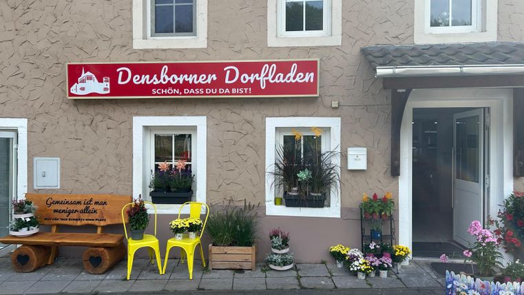 Ein kleiner Dorfladen mit dem Namen "Densborner Dorfladen" an einer hellen Fassade. Vor dem Laden stehen bunte Blumen und eine Holzbank zum Sitzen.