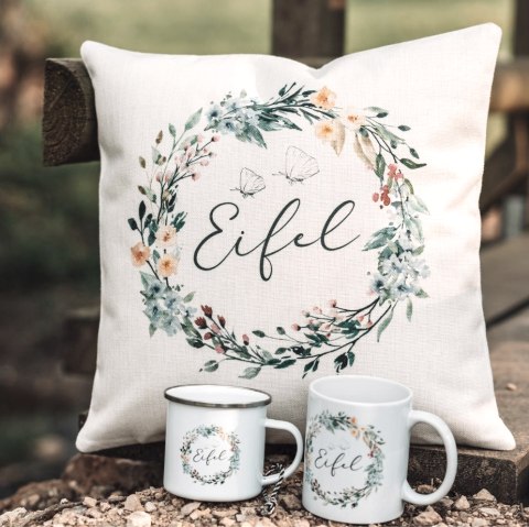 Handgemachtes Stoffkissen mit Blumemuster und Schriftzug "Eifel" in der Mitte auf einem Holzsteg. Davor zwei Tassen mit dem gleichen Design von Mitgebracht.