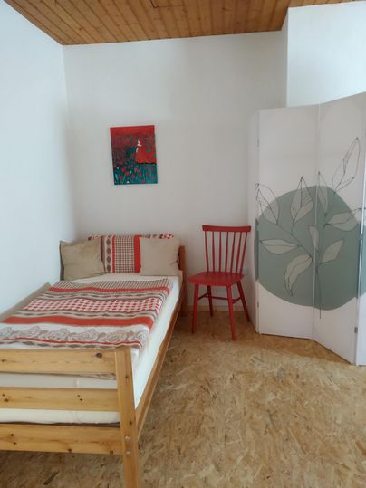 Une chambre simple avec un lit, une chaise rouge et un paravent. Un tableau coloré est accroché au mur.