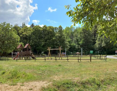 Playground with wooden houses, swings, and climbing frame, surrounded by trees and grass, under blue sky with clouds.