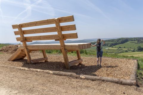 Eine riesige Holzbank steht in einer ländlichen Landschaft. Eine Person steht daneben und schaut in die Ferne.