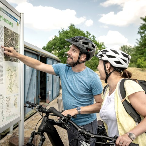 Two cyclists with helmets study an information board on the Kyll cycle path. They smile and point to the map. In the background are trees and a blue container., &copy; Eifel Tourismus GmbH, Dominik Ketz