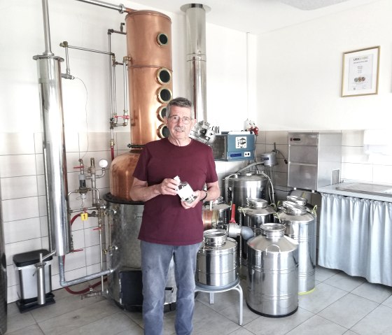 Ein Mann vor einer Destilliermaschine mit einer Flasche Gin in der Hand.