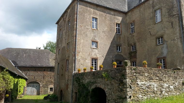 Die Aufnahme zeigt eine alte Burg oder ein Herrenhaus mit einem Steingebäude im Vordergrund und einem weiteren Gebäude aus Stein und Holz im Hintergrund. Die Gebäude sind von Grünflächen umgeben.