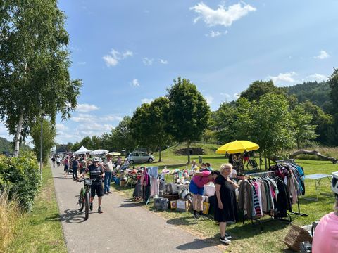 Ein Flohmarkt am Wegesrand mit Ständen, die Kleidung und andere Waren anbieten. Menschen schlendern vorbei und genießen das schöne Wetter.