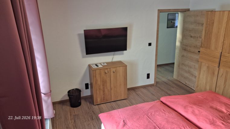 Ein modernes Schlafzimmer mit einem Doppelbett und einem Fernseher an der Wand. Neben dem Bett steht ein Holzmöbelstück und ein Abfalleimer ist sichtbar.