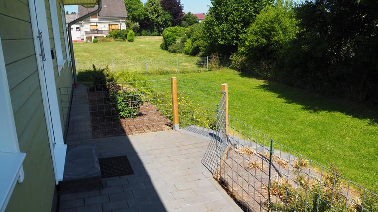 Eine Terrasse mit Pflastersteinen und einem Sichtschutz aus Gitter. Im Hintergrund sieht man einen grünen Garten mit Bäumen und einem Einfamilienhaus.