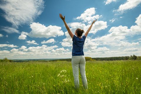 Eine Frau steht auf einer hochgewachsenen Wiese unter blauem Himmel, schaut in die Ferne und hält ihre Arm weit in die Luft.