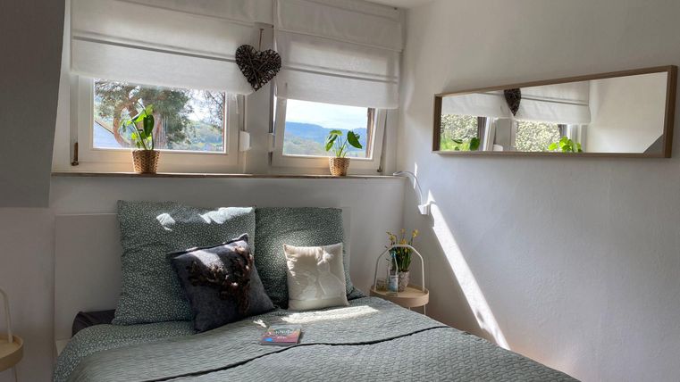 Ein gemütliches Schlafzimmer mit einem Bett und mehreren Kissen. Die Fenster sind hell und bringen viel Tageslicht herein.