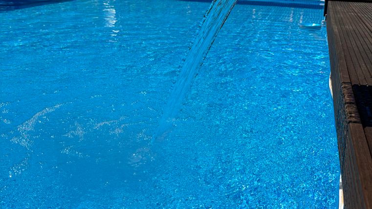 Ein Pool mit klarem, blauem Wasser, aus dem ein Wasserstrahl fließt. Im Hintergrund sind entspannende Liegen und grüne Bäume zu sehen.