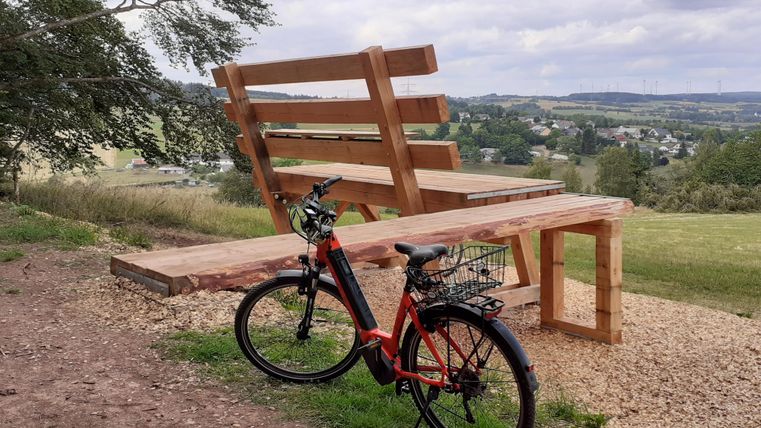 Eine übergroße Holzbank steht auf einem Hügel mit Blick auf eine ländliche Landschaft. Ein Fahrrad lehnt an der Bank.