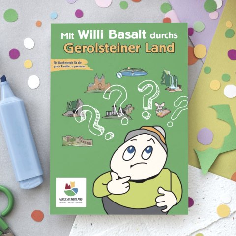 Titelseite  des R&auml;tselhefts "Mit Willi Basalt durch das Gerolsteiner Land" auf einem Tisch voller Mal- und Bastelutensilien und bunter Bl&auml;tter.